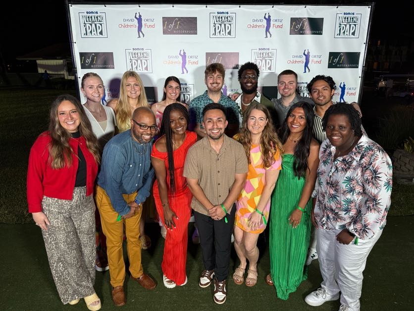 A group of people poses for a photo in front of a backdrop featuring event sponsors. They are dressed in various casual and semi-formal attire.