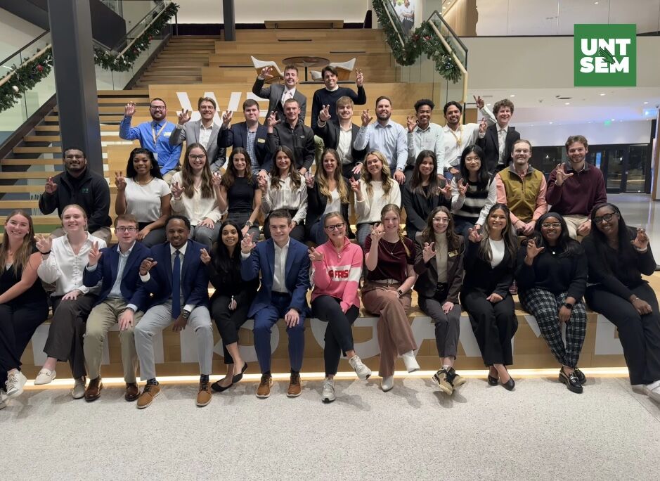 A large group of students and faculty are posing for a photo while sitting and standing on tiered wooden steps. They are all looking at the camera and making the UNT Eagle Claw hand gesture.
