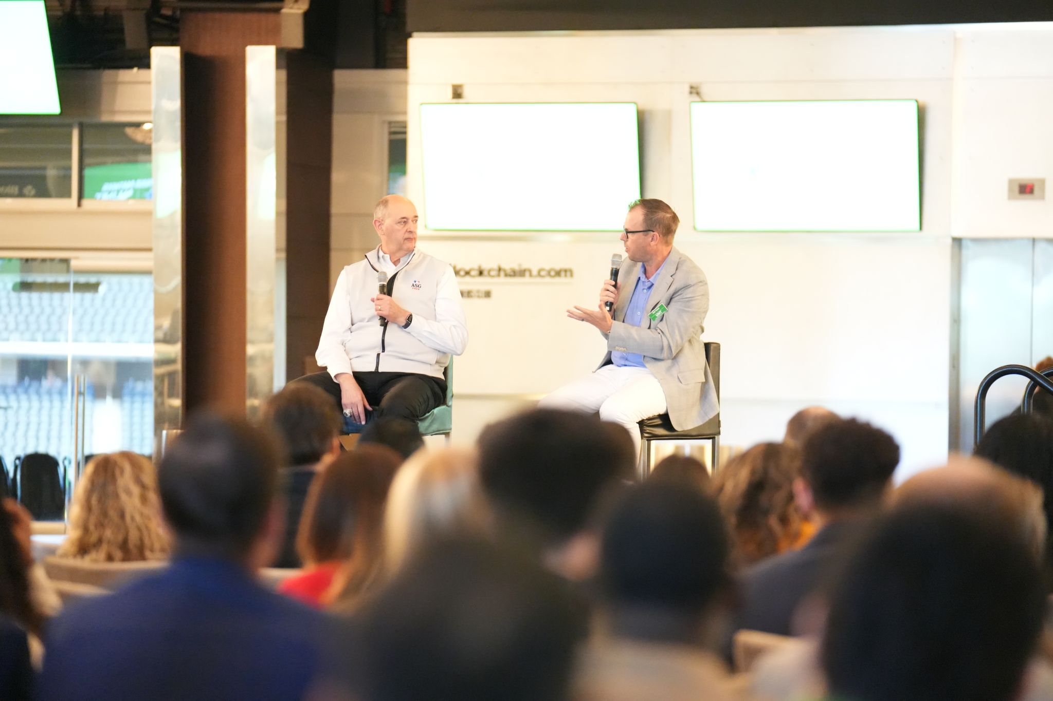 Two men are sitting on chairs, each holding a microphone and speaking to an audience. They appear to be participating in a panel discussion or presentation.
