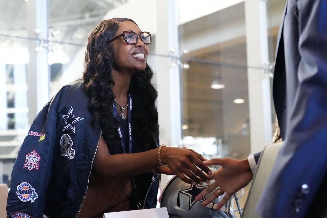 Dexia Smith, wearing a Dallas Cowboys jacket, shakes hands with a man in a suit at a table with a Cowboys helmet and a name card.