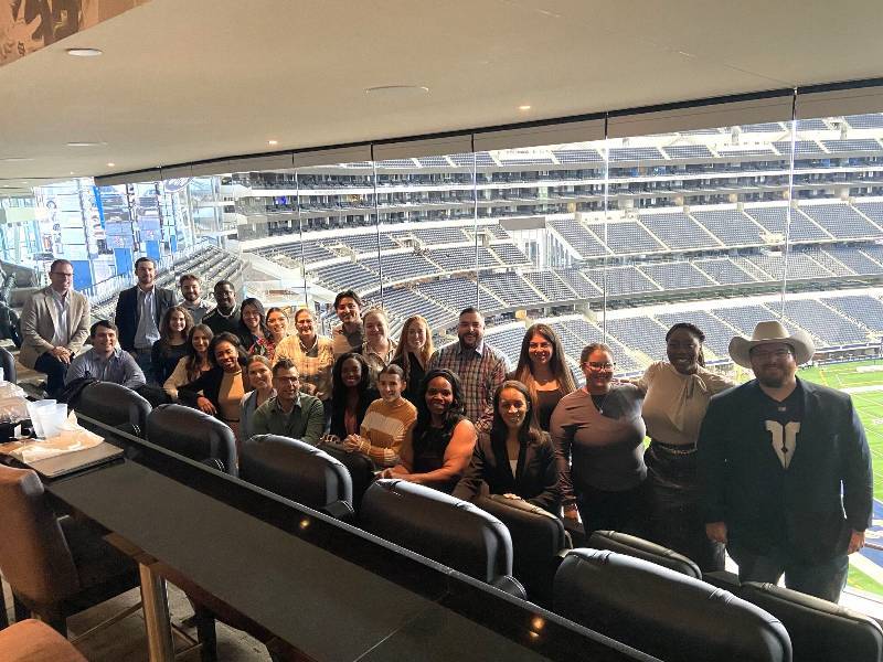 A large group of people are posing for a photo in a stadium suite. The stadium seats are visible in the background.