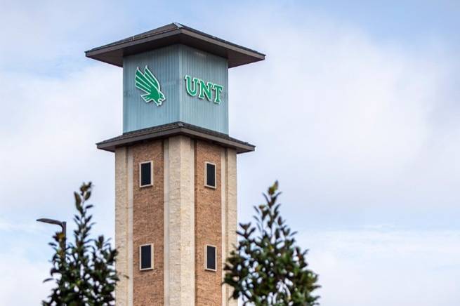 A tall tower with the UNT logo stands against a cloudy sky, partially obscured by greenery. The tower has brick and stone construction with multiple windows.