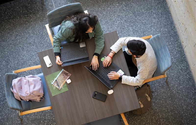Two individuals are working together on laptops at a table, with books, phones, and backpacks nearby. The scene is captured from an overhead perspective.