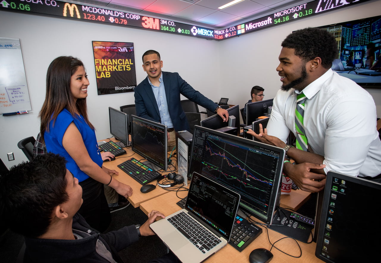 A group of young adults are gathered around computer monitors displaying financial data in a lab. They appear to be collaborating and discussing the information on the screens.