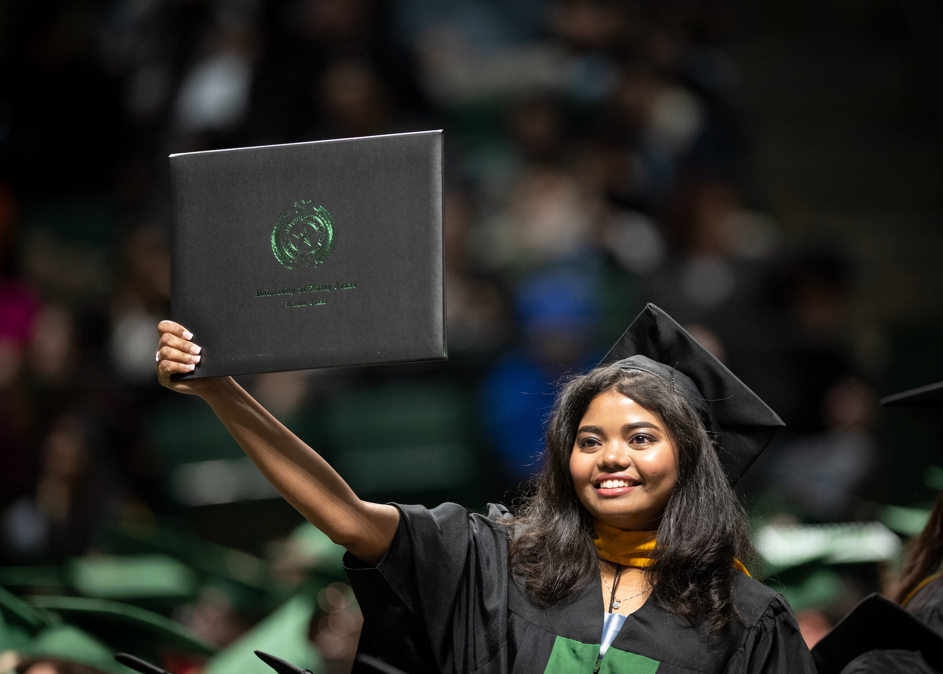 A smiling graduate in a cap and gown holds up a diploma sleeve. The diploma sleeve displays the University of North Texas logo.