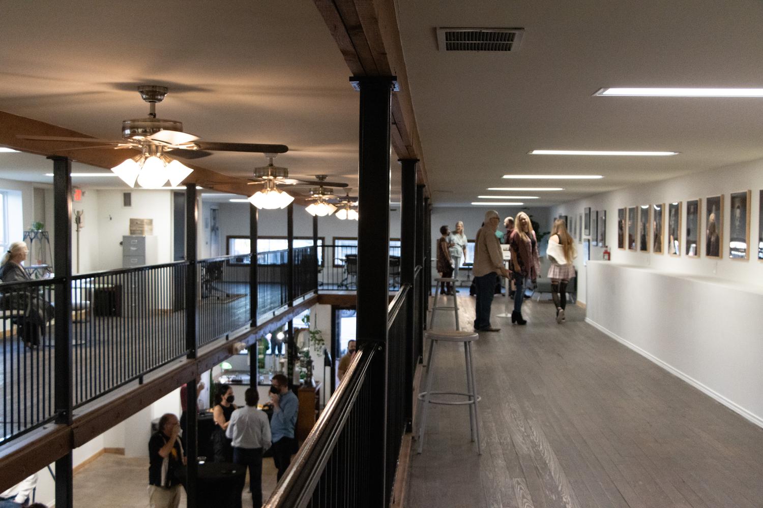 People are gathered at a multi-level venue, with some on a balcony overlooking a lower floor. Another group of people are observing framed art displayed on a wall in a long hallway.