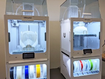 Two Ultimaker 3D printers are shown with white prints inside and spools of filament in the storage compartments.