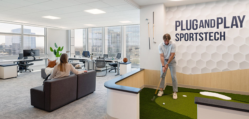 A man plays miniature golf in a modern office space with a woman sitting on a couch in the background. The wall behind the golf area has the text "PLUG AND PLAY SPORTS TECH" displayed.