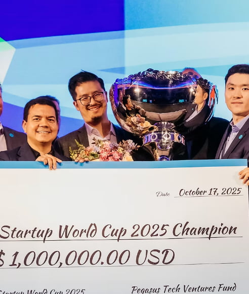 A group of men are celebrating winning the Startup World Cup 2025, holding a trophy and a giant check for $1,000,000.00 USD.